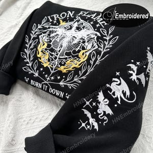 K&ouml;nnte beinhalten: Schwarzer Sweatshirt mit wei&szlig;er und goldener Stickerei. Die Vorderseite zeigt ein Design mit dem Text "IRON FLAME" und "BURN IT DOWN". Die &Auml;rmel zeigen ein sich wiederholendes Muster aus Drachen und Sternen.