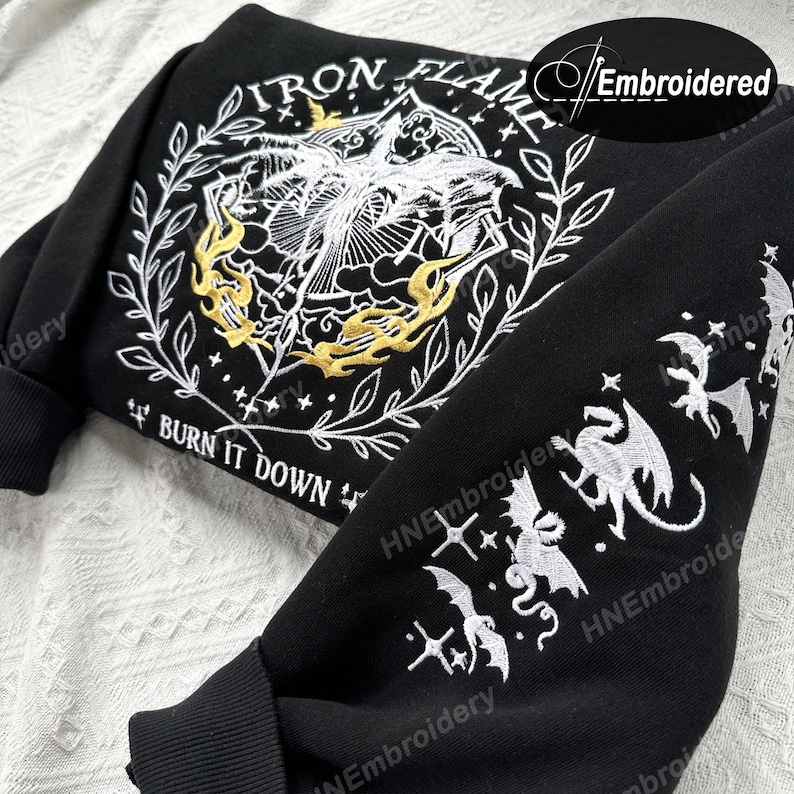 K&ouml;nnte beinhalten: Schwarzer Sweatshirt mit wei&szlig;er und goldener Stickerei. Die Vorderseite zeigt ein Design mit dem Text "IRON FLAME" und "BURN IT DOWN". Die &Auml;rmel zeigen ein sich wiederholendes Muster aus Drachen und Sternen.