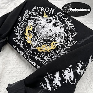 K&ouml;nnte beinhalten: Schwarzer Sweatshirt mit wei&szlig;er und goldener Stickerei. Das Design zeigt einen stilisierten Vogel mit Flammen und dem Text "IRON FLAME" und "BURN IT DOWN".