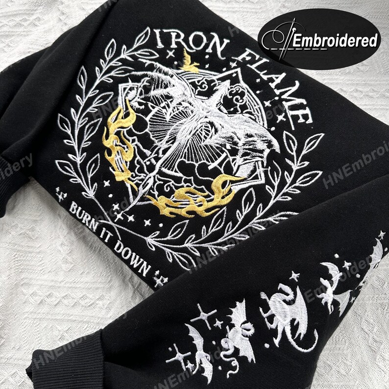 K&ouml;nnte beinhalten: Schwarzer Sweatshirt mit wei&szlig;er und goldener Stickerei. Das Design zeigt einen stilisierten Vogel mit Flammen und dem Text "IRON FLAME" und "BURN IT DOWN".