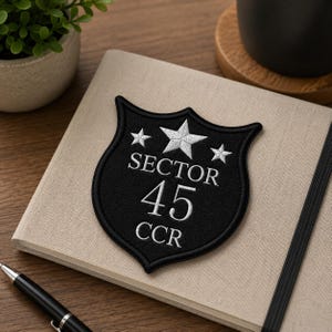 May include: A black embroidered patch in a shield shape, with the text "SECTOR 45 CCR" in white, and three white stars. The patch is on a beige notebook, with a pen and a small potted plant visible.