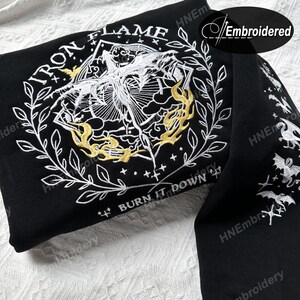 K&ouml;nnte beinhalten: Schwarzer Hoodie mit wei&szlig;er und goldener Stickerei. Die Stickerei zeigt eine gefl&uuml;gelte Kreatur mit den Worten "Iron Flame" und "Burn It Down" darum herum.