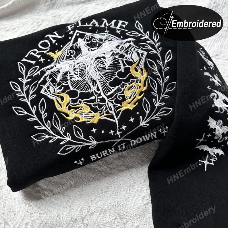 K&ouml;nnte beinhalten: Schwarzer Hoodie mit wei&szlig;er und goldener Stickerei. Die Stickerei zeigt eine gefl&uuml;gelte Kreatur mit den Worten "Iron Flame" und "Burn It Down" darum herum.