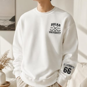 Op de afbeelding: Een witte crewneck sweatshirt met zwarte tekst en afbeeldingen. De voorkant toont "BRIAR UNIVERSITY" met gekruiste hockeysticks en het jaar "2015". De rechter mouw heeft "DI LAURENTIS 66" in zwart.