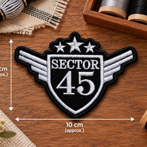 May include: A black and white embroidered patch featuring a shield design with the text SECTOR 45, three stars above, and wings on the sides. The patch measures approximately 2.95 inches in height and 3.94 inches in width, as indicated by arrows.