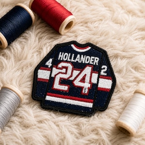 Könnte beinhalten: Ein marineblaues Hockey-Trikot-Patch mit dem Namen "Hollander" und der Nummer "24" in Weiß. Der Patch hat rote und weiße Streifen und ist von einem schwarzen Rand umgeben. Mehrere Garnrollen in verschiedenen Farben sind um den Patch herum verstreut.