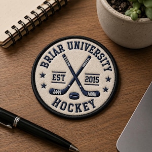 May include: A circular patch with the words "Briar University Hockey" in navy blue. The patch features crossed hockey sticks, a puck, and the text "EST 2015". The patch is cream-coloured with a navy blue border and stars.