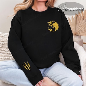 May include: Black sweatshirt with a gold embroidered Witcher medallion on the chest and claw marks on the left sleeve. The sweatshirt is a crew neck style and is made of a soft material. The word "Embroidered" is in the upper right corner.