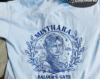 Op de afbeelding: Lichtblauw T-shirt met een geborduurd ontwerp. Het ontwerp bevat de naam "Minthara" in een cirkelvormig patroon, een portret van een vrouw en de tekst "Baldur's Gate". Het ontwerp is in blauwtinten.