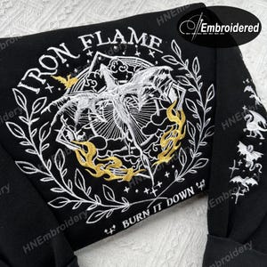 K&ouml;nnte beinhalten: Schwarzer Sweatshirt mit wei&szlig;er und goldener Stickerei. Das Design zeigt eine gefl&uuml;gelte Kreatur mit Flammen und dem Text "IRON FLAME" und "BURN IT DOWN".