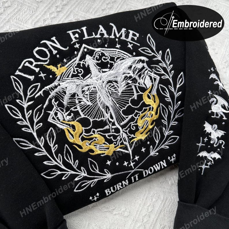 K&ouml;nnte beinhalten: Schwarzer Sweatshirt mit wei&szlig;er und goldener Stickerei. Das Design zeigt eine gefl&uuml;gelte Kreatur mit Flammen und dem Text "IRON FLAME" und "BURN IT DOWN".