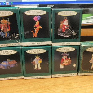 May include: Eight small green boxes with white lettering, each containing a different Christmas ornament. The ornaments include a Santa Claus on a snowmobile, a Winnie the Pooh and Tigger ornament, a blue truck, a Santa Claus with a beard, a drummer boy, and a ballerina.