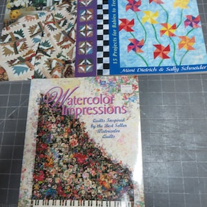 May include: Three quilting books with colorful covers. The first book is titled "Fabulous Quilts from Favorite Patterns" and features a quilt with a variety of patterns. The second book is titled "Growing Up With Quilts" and features a quilt with a colorful floral design. The third book is titled "Watercolor Impressions" and features a quilt with a floral design on a piano.