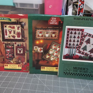 May include: Three Christmas quilt patterns featuring a gathering of angels, a collection of Santas, and whispering pines. Each pattern includes the text "Mumm's the Word".