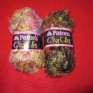 May include: Two skeins of Patons Cha Cha yarn. One skein is pink, gold, and purple. The other skein is brown, gold, and black.