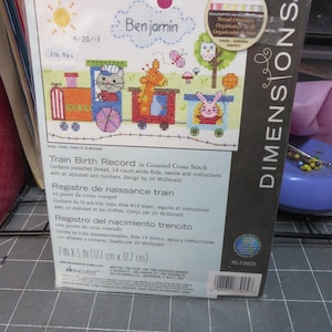 May include: A Dimensions counted cross-stitch kit featuring a colorful train with animal passengers. The kit includes pre-sorted thread, aida cloth, needle, and instructions. The package is 7 inches by 5 inches (17.7 cm x 12.7 cm) and includes the name "Benjamin".
