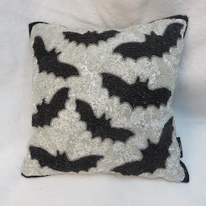 May include: Square decorative pillow with a pattern of black bats on a silver beaded background. The pillow has a black border and is likely used for home decor, especially for Halloween.
