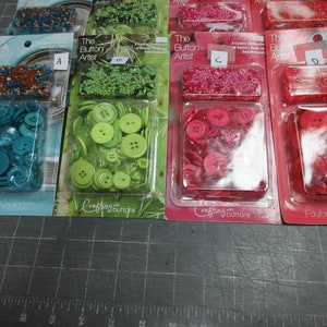May include: Four packages of assorted buttons in different colors. The packages are labeled "The Button Artist" and feature a variety of buttons in green, red, and blue.