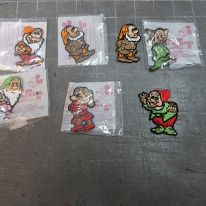 May include: A collection of seven embroidered patches featuring characters from a classic animated film. Each patch depicts a different dwarf in vibrant colors, packaged in individual clear plastic sleeves.
