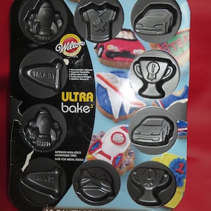 May include: A 12-cavity cookie pan with a blue and white background. The pan is designed for making boy-themed cookies. The cavities are shaped like rockets, cars, trophies, and t-shirts. The pan is made of aluminum and has a non-stick coating. The text "ULTRA bake" is printed on the pan in black letters. The text "12 CAVITY BOY COOKIE PAN" is printed on the pan in black letters. The text "Recipe on Back" is printed on the pan in black letters.
