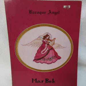 May include: A cross-stitch pattern titled "Baroque Angel" by Mar Bek. The cover is a deep red with the title in elegant script. The image features an angel in a pink dress playing a violin, surrounded by a gold oval frame.