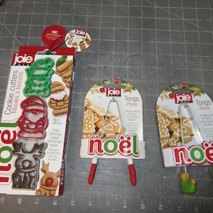 May include: Three Christmas-themed kitchen tools: a set of three cookie cutters shaped like Santa, a reindeer, and a Christmas tree, a pair of red tongs with the word "noël" printed on them, and a pair of green tongs with the word "noël" printed on them.