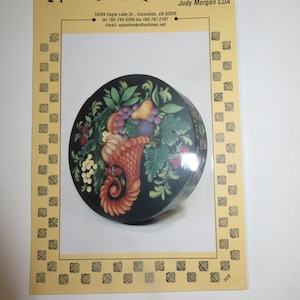 May include: A round wooden box with a painted design of a cornucopia overflowing with fruit, grapes, and leaves. The box is painted on a black background with gold accents. The text "Apple Cheeks Publications" is printed at the top of the image.