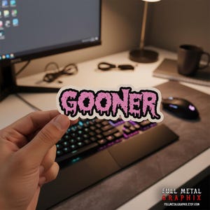 May include: A white sticker with the word "GOONER" in dripping pink letters. The sticker is held in front of a keyboard and computer monitor. The sticker is from Full Metal Graphix.
