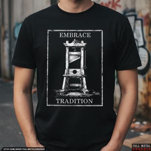 May include: A black short-sleeve t-shirt featuring a white graphic print of a guillotine centered within a rectangular frame. The text EMBRACE is printed above the guillotine and TRADITION is printed below it in a bold, white, serif font.