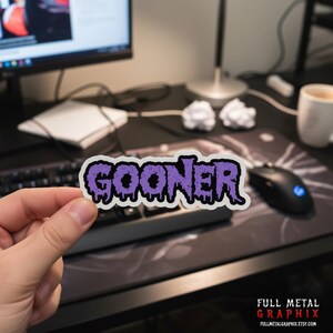 May include: A rectangular sticker with the word "GOONER" in dripping purple letters outlined in black and white. The sticker is held in front of a computer setup with a keyboard, mouse, and monitor.