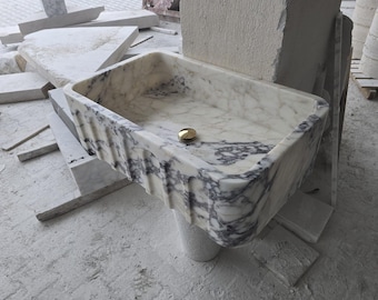 Puede incluir: Un lavabo de mármol blanco con vetas grises. El lavabo tiene una forma rectangular con bordes redondeados y un desagüe en el centro. El lavabo está colocado sobre un pedestal de mármol.