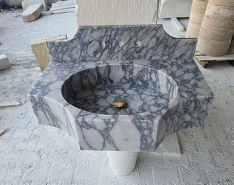 Op de afbeelding: Een witte marmeren wastafel met een paars adermotief. De wastafel heeft een afgerond, ovaalvormig bassin en een decoratieve achterwand met drie gaten. De wastafel is gemonteerd op een witte voet.