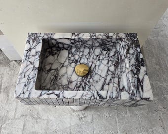 May include: Rectangular marble sink with a gold-colored drain cover. The sink features a white and black veined pattern. The edges are made of small, rectangular tiles. A modern bathroom fixture.