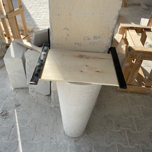 May include: A close-up of a stone column being measured with a wooden board and metal brackets. The column is made of rough, textured stone and is being worked on in a construction setting.