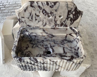 Puede incluir: Un lavabo de mármol blanco con vetas moradas. El lavabo tiene un borde decorativo acanalado y un salpicadero con tres agujeros. El lavabo es independiente y parece estar hecho de piedra natural.