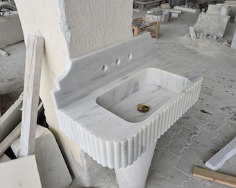 Puede incluir: Un lavabo de baño de mármol blanco con una base acanalada y un solo agujero para grifo. El lavabo está montado en una pared y tiene un salpicadero de mármol blanco.
