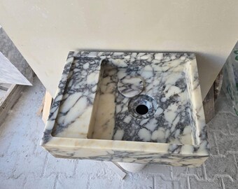 May include: Rectangular marble sink with a white and gray veined pattern. The sink has a rectangular basin, a drain hole, and a small, circular overflow. The sink is made of a natural stone material.