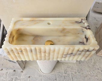 Op de afbeelding: Een rechthoekige, crèmekleurige marmeren wastafel met een gegroefde rand en een goudkleurige afvoer. De wastafel heeft een rechthoekige bak en een klein gat voor de kraan. Het marmer heeft bruine en paarse aders.