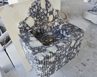 May include: A marble sink with a speckled pattern of grey and white. The sink has a rectangular basin and a decorative back with a curved top. A gold-coloured drain is visible.