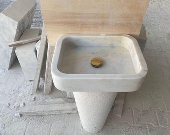 May include: A white marble bathroom sink with a gold drain stopper. The sink is mounted on a pedestal made of the same material.