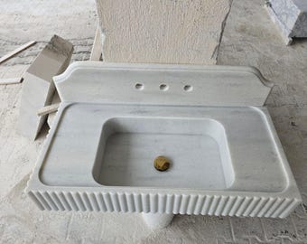 Puede incluir: Lavabo de baño de mármol blanco con base acanalada y un solo desagüe. El lavabo tiene un borde superior decorativo y tres agujeros para accesorios.