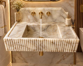 Può includere: Un lavabo in marmo a parete con design frontale scanalato e rubinetto in oro lucido. Il lavabo include due valvole a manopola a croce dorate, uno scarico dorato abbinato e un paraschizzi in marmo. L'acqua scorre dal rubinetto centrale.