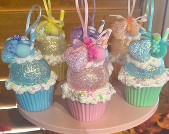 May include: A collection of glittery cupcake ornaments in various pastel colors, including blue, pink, yellow, and orange. Each ornament features a ribbon loop for hanging and decorative details like bows and faux whipped cream.