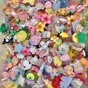 May include: A collection of colorful resin charms in various shapes, including flowers, animals, bows, and stars. The charms are in shades of pink, yellow, blue, and green, with some featuring glitter and other decorative elements. These are ideal for crafting and jewelry making.