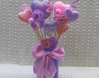 Può includere: Un vaso viola con un fiocco rosa contiene un bouquet di cuori glitterati rosa e viola con fiori rosa e viola.