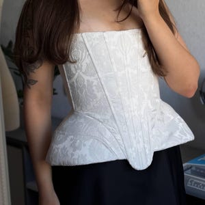 May include: A strapless white corset top featuring a subtle tonal damask floral pattern, vertical boning channels, and a structured peplum hem that flares at the waist. The garment is crafted from a textured fabric with a matte finish.