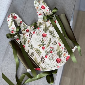May include: A corset-style top featuring a cream-colored fabric with a botanical print of red strawberries, green ferns, and foliage. The garment includes vertical stitching, wide shoulder straps, and long olive green satin ribbons for tying.