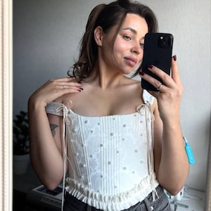 Può includere: Un top a corsetto bianco con abbellimenti di perle e un motivo floreale. Il top presenta spalline scoperte con dettagli a fiocco e un orlo a balze. Il corsetto è abbinato a jeans di denim grigio scuro.