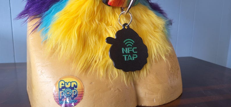 May include: A black, cloud-shaped NFC tag with green text reading NFC TAP and a Wi-Fi symbol, attached to a rainbow-colored faux fur collar. A circular sticker with the text Fur Pop and a paw print icon is visible on the tan mannequin base.