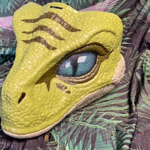 Puede incluir: Una hucha con forma de cabeza de dinosaurio amarilla con un ojo detallado y piel texturizada. El ojo es azul con una pupila oscura. La hucha est&aacute; sobre una tela con un estampado de hojas tropicales.